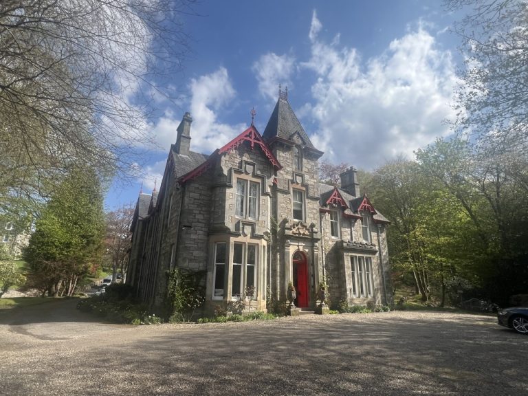 Grey stone victorian house stands with a vibrant red front door and matching roof skirting